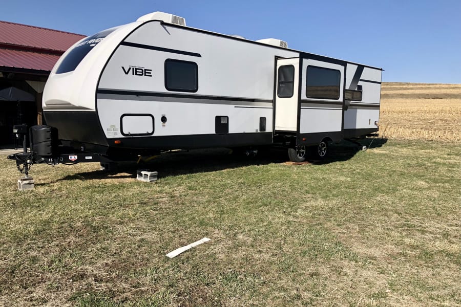 This Forest River Vibe travel trailer, ready for an RV rental adventure, is set in a grassy field beside a barn and farmland.