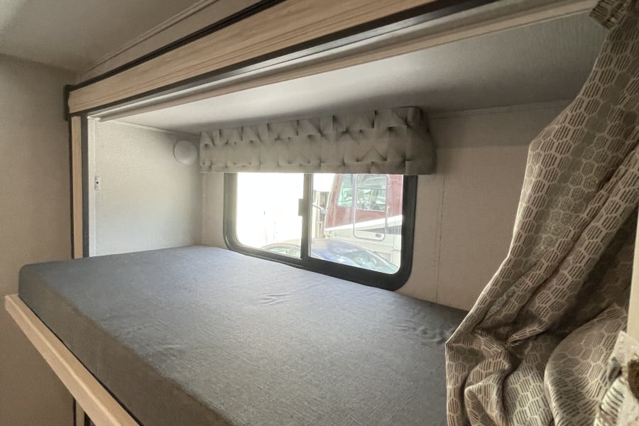 RV rental bedroom interior featuring a grey mattress, window overlooking vehicles, and patterned drapes, showcasing comfort.