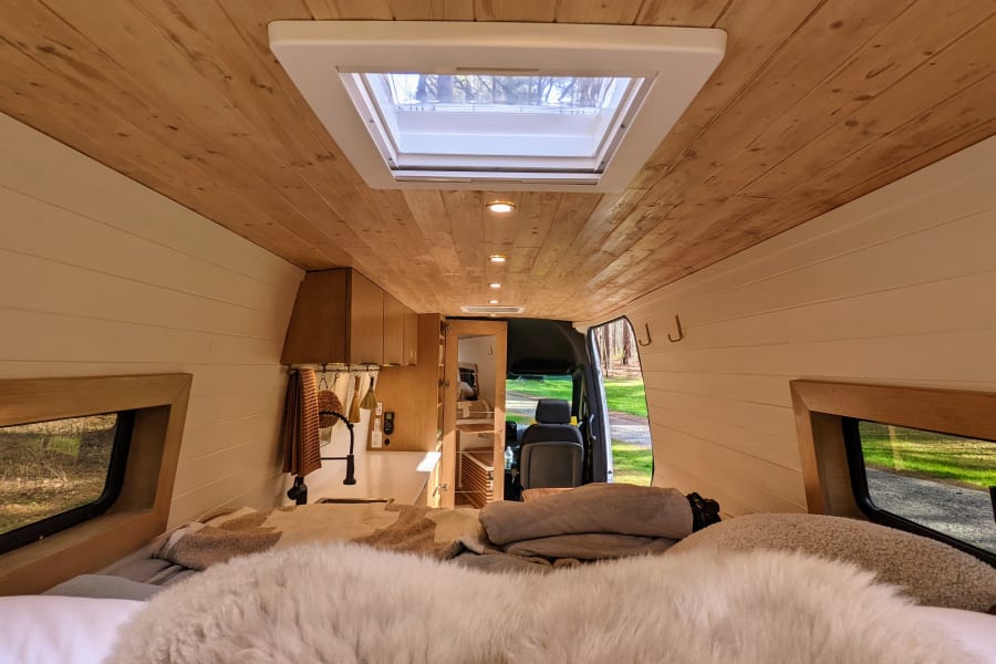 Cozy rv rental interior: view from the bed with fluffy blankets, showcasing a wood ceiling, skylight, and galley kitchen.