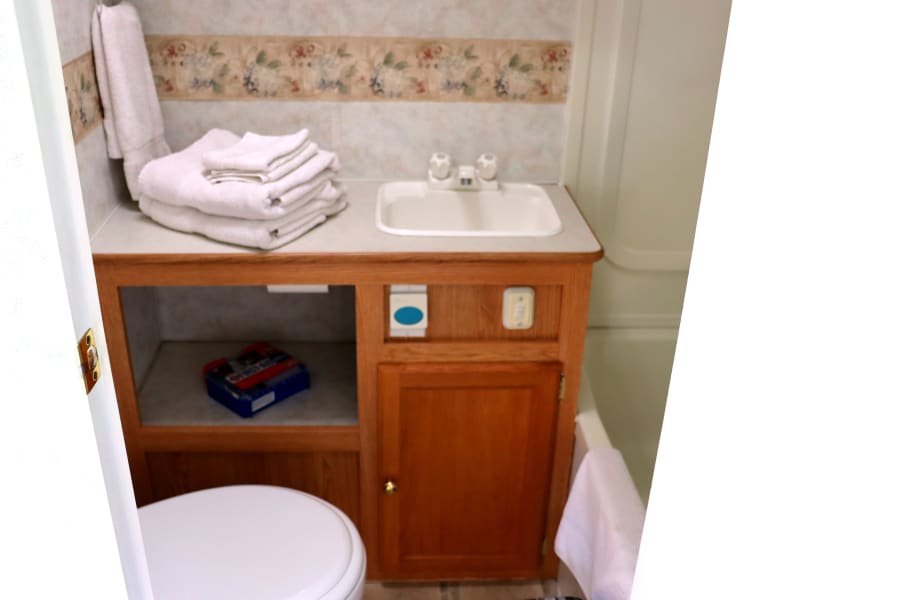 Compact RV rental bathroom featuring a wooden vanity, sink, toilet, and stacked white towels.