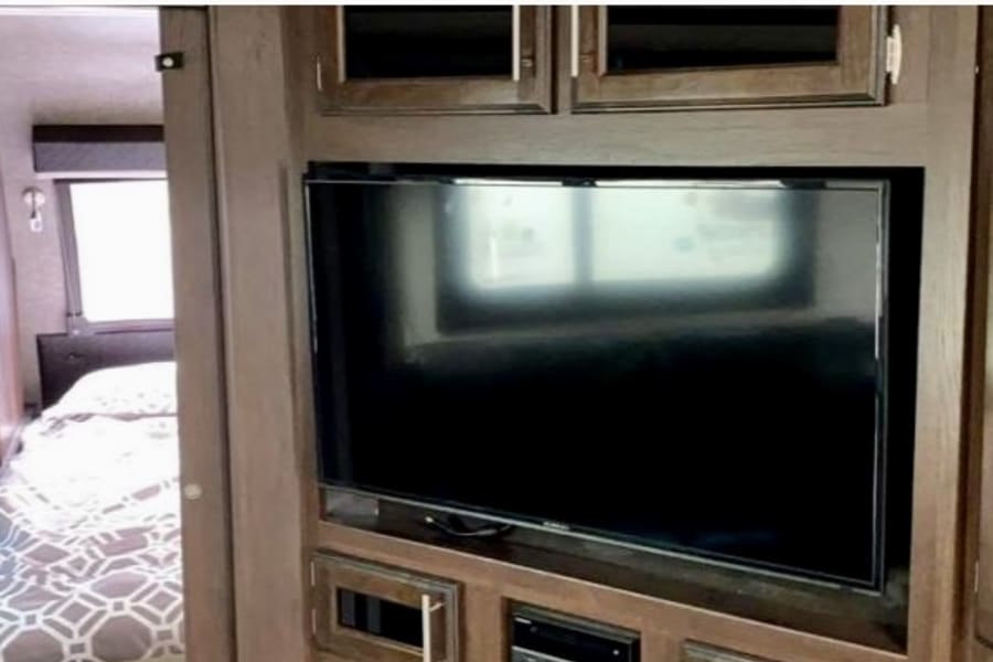 RV rental interior featuring a large mounted TV within dark wood cabinetry, with a cozy bedroom visible through a doorway.