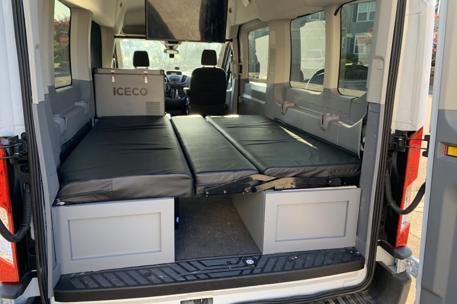 Rear view of an **RV rental** camper van interior with an ICECO fridge, two black foldable beds, and a mounted TV.