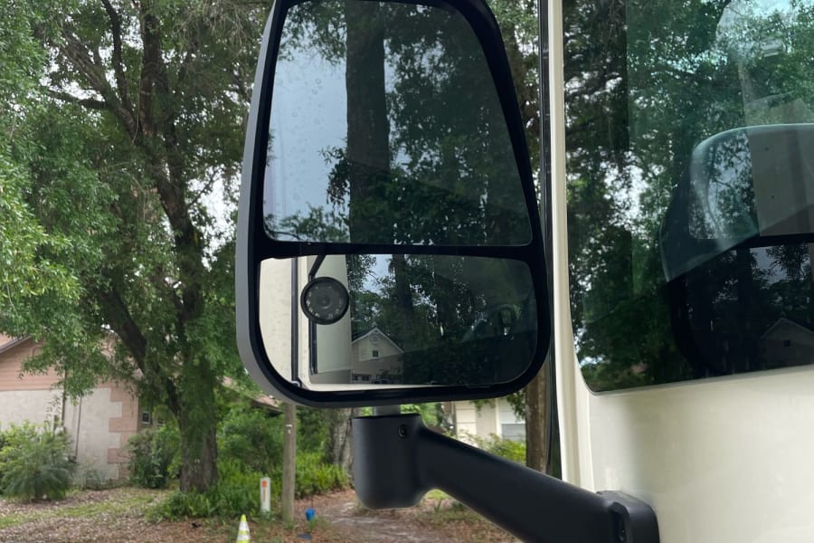 Close-up of an RV rental's side mirror, reflecting a residential street, trees, and distant houses.