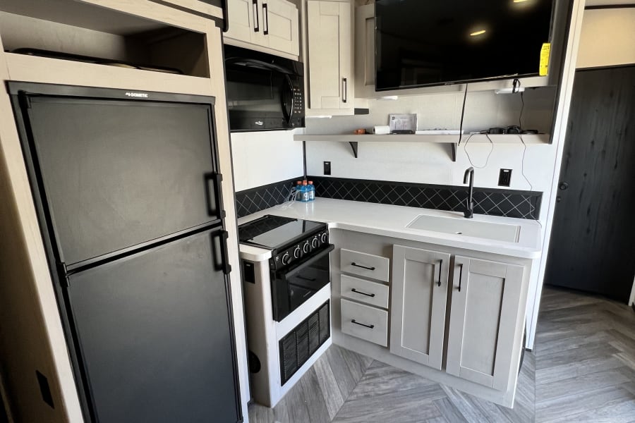 Modern RV rental kitchen with fridge, stove, sink, and TV. Note the herringbone floor and utility tie-downs.