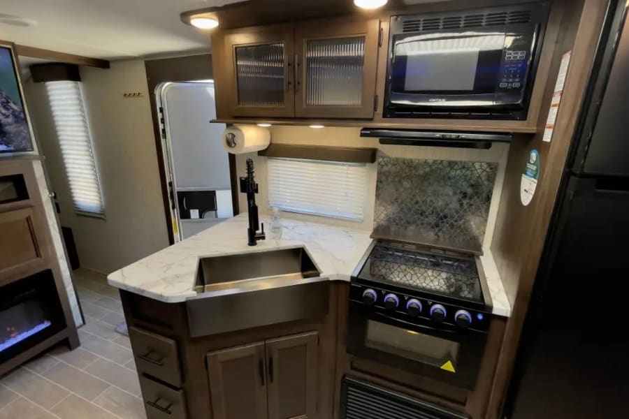 Spacious rv rental kitchen featuring microwave, stove, sink, & fridge. Living area has a TV, fireplace, and modern decor.