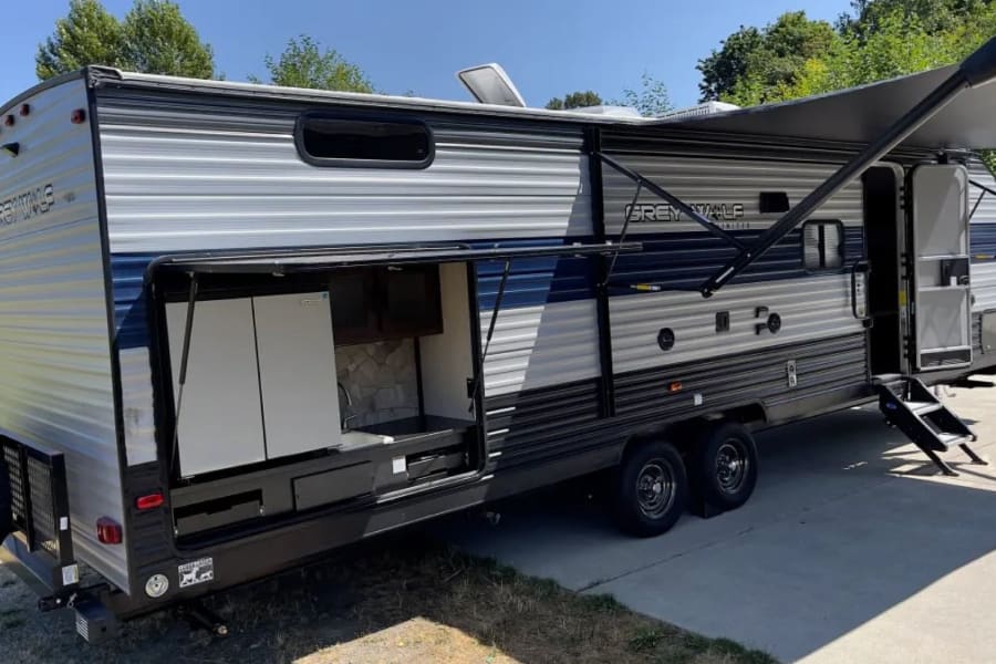 Sunny day view of an rv rental: a silver and blue travel trailer with its outdoor kitchen open, awning extended, and entry steps down.
