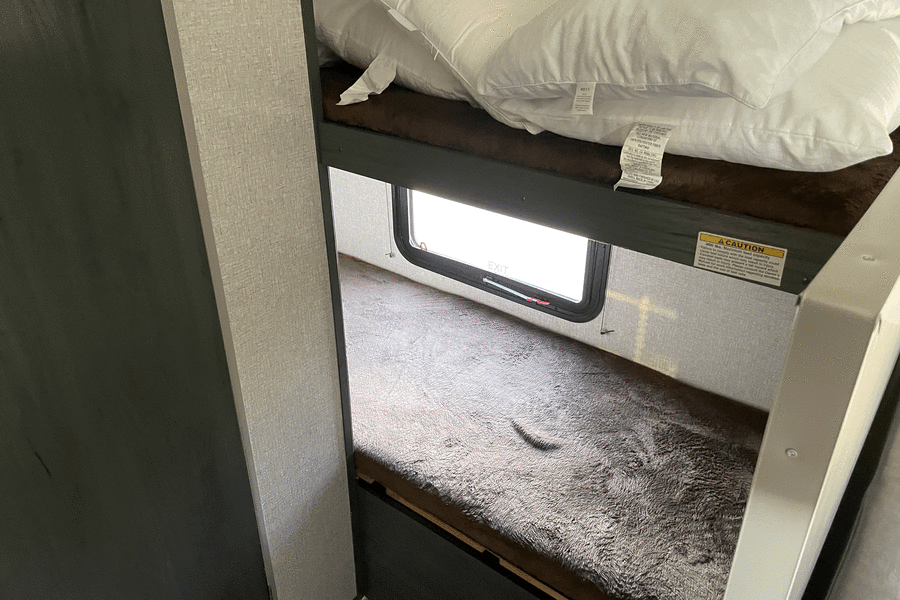 **RV rental** bunk beds: upper with pillows, lower with plush blanket, featuring an exit window and safety label.