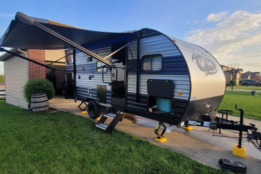 A modern travel trailer, ready for your rv rental, is parked in a driveway with its awning extended on a sunny day.