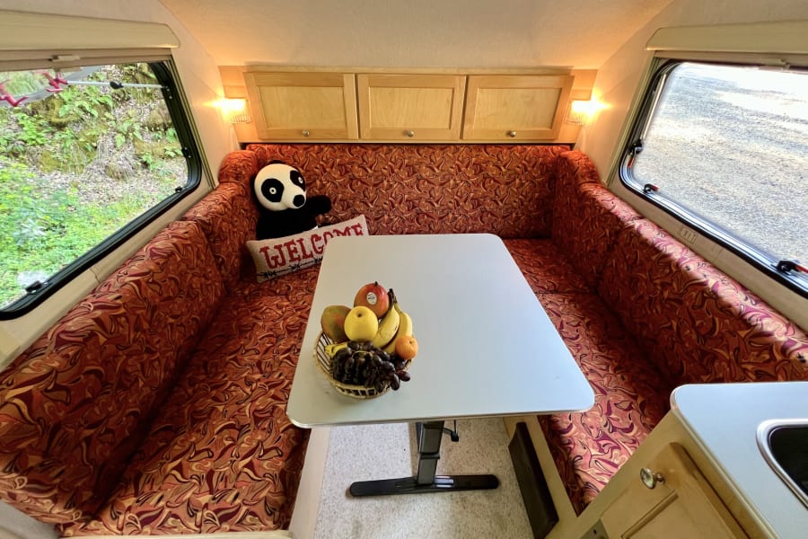 Inside a comfortable rv rental: a well-lit dinette, fruit basket, plush panda, and scenic window views.