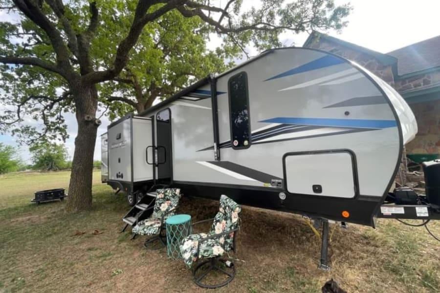 Modern grey travel trailer RV rental by a tree, with outdoor seating & extended slide-out for campsite comfort.