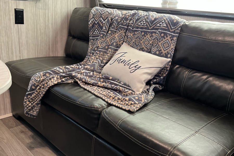 Relax in this RV rental's comfortable interior! Features a dark sofa with a patterned blanket and pillow.