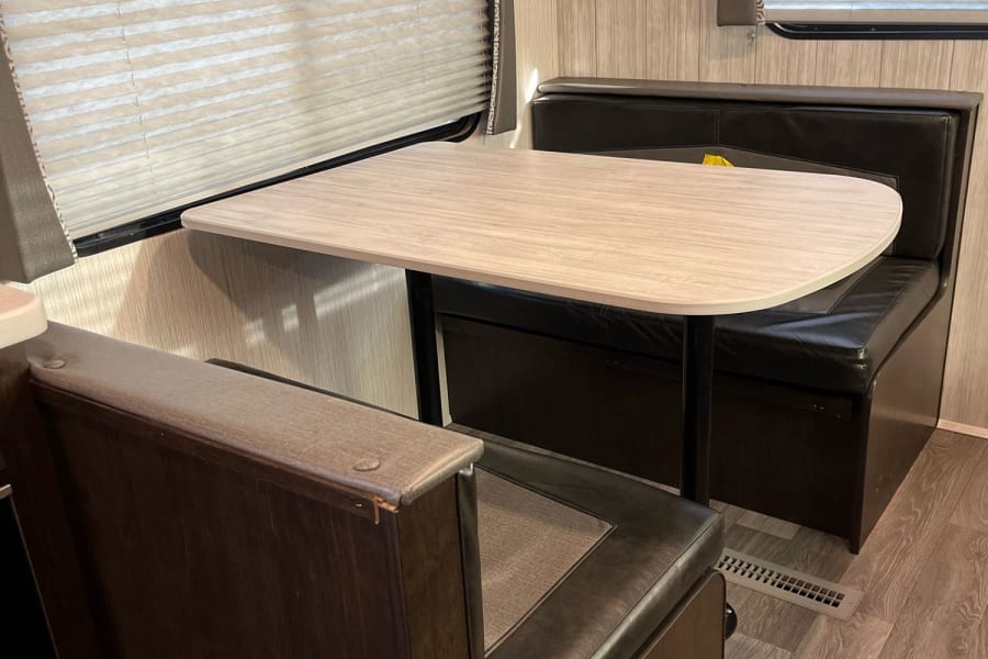 RV rental dinette with a light wood table, black benches, and pleated blinds on the windows.