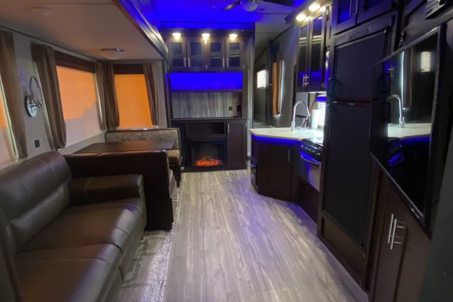 Experience a spacious RV rental interior, showcasing a living area, kitchen, and dining booth with modern blue lighting and a cozy fireplace.