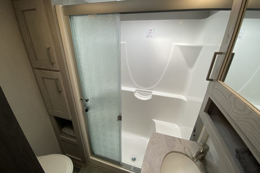 Compact RV rental bathroom: features a white shower with frosted glass slider, toilet, and a vanity with sink and storage.