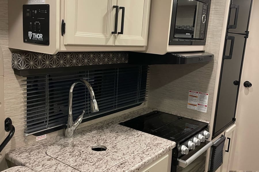 Compact yet functional RV rental kitchen: modern stove, microwave, large sink, ample counter space, cream cabinets, and full-size fridge.