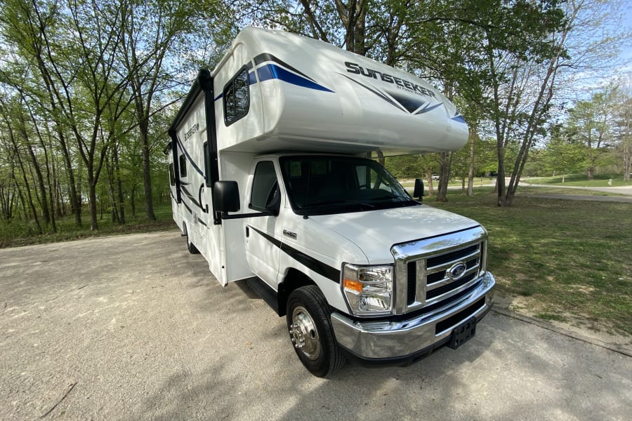 White Sunseeker RV rental parked on a paved area, surrounded by green trees under a sunny sky.