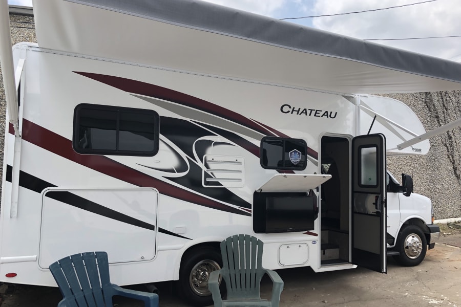 A white Chateau RV rental is shown with its extended awning, an outdoor TV in an open compartment, and two patio chairs.