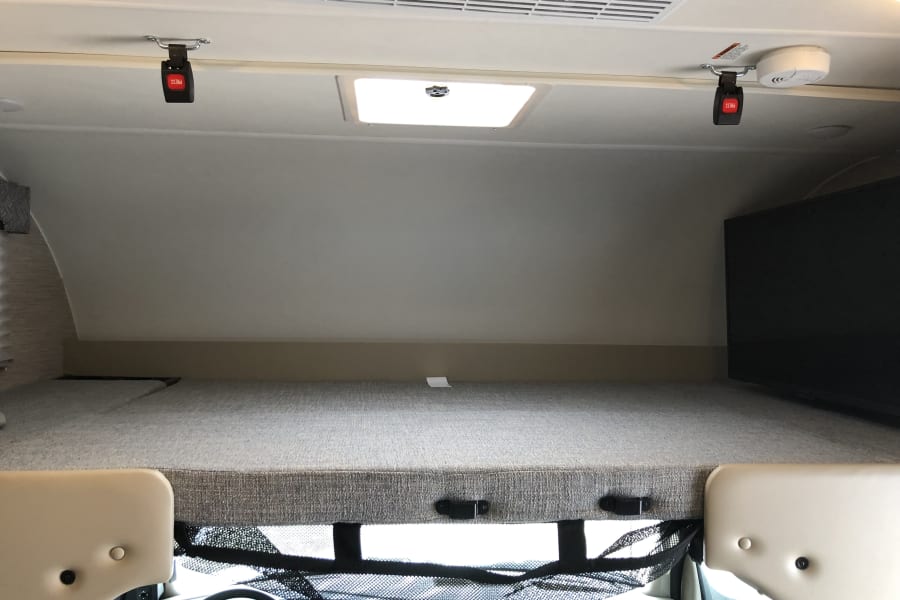 RV rental interior: Cab-over bunk bed with TV, safety net, overhead AC, skylight, smoke detector, and seat belt buckles.