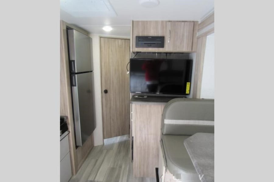 Inside a modern RV rental: stainless steel fridge, dining booth, TV, and kitchen. Light wood tones & flooring.