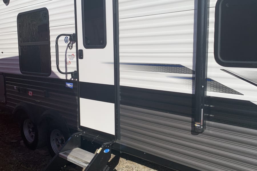 A modern RV rental's side, featuring an open entry door with steps, windows, and an awning system. Parked outside on gravel.