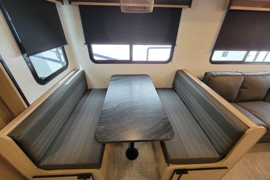 Bright RV rental dinette area featuring dark grey cushioned benches, a patterned table, and windows with blinds.