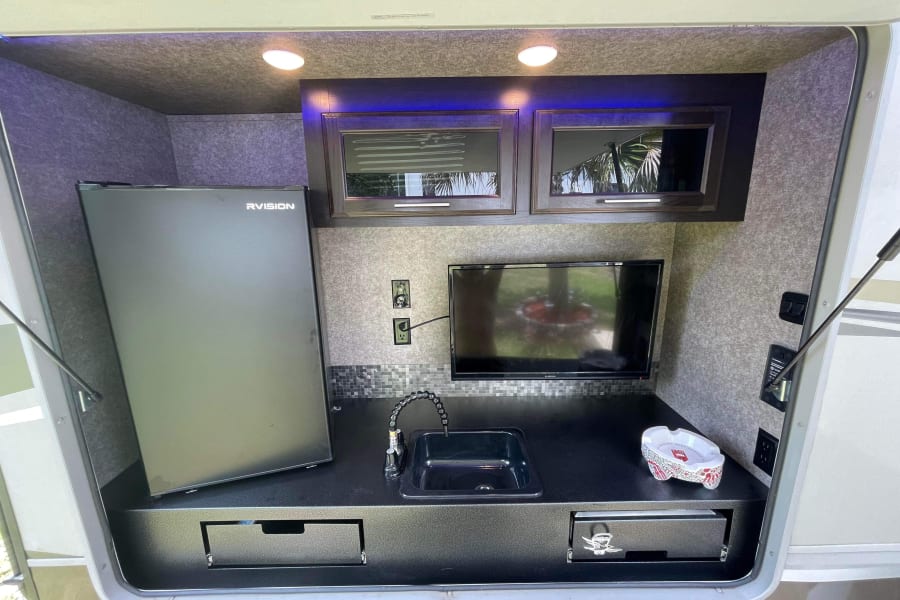 This RV rental features a well-equipped outdoor kitchen: mini-fridge, sink, TV, cabinets & drawers for camping convenience.