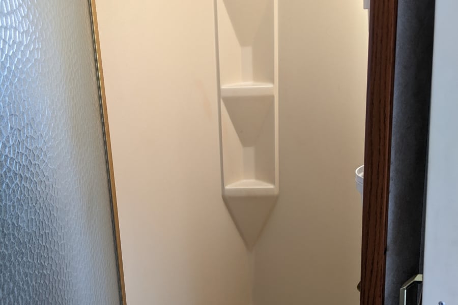 This RV rental features a compact beige corner shower unit with built-in shelves, textured glass door, and dark shower mat.