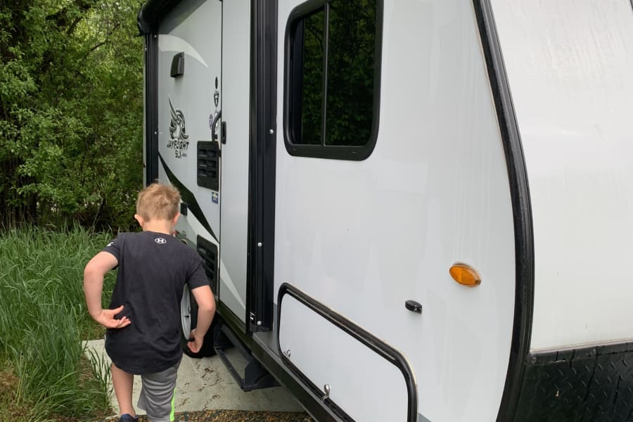 Child steps into an RV rental, a travel trailer with a black awning, amidst green grass and trees.
