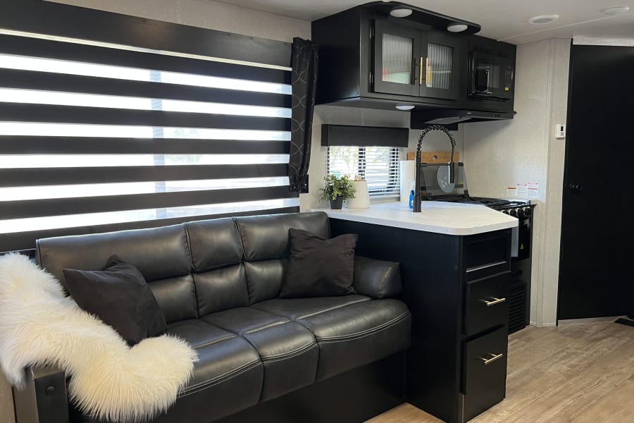 Cozy RV rental interior: black leather sofa, modern kitchen with white counters, and large striped blinds.