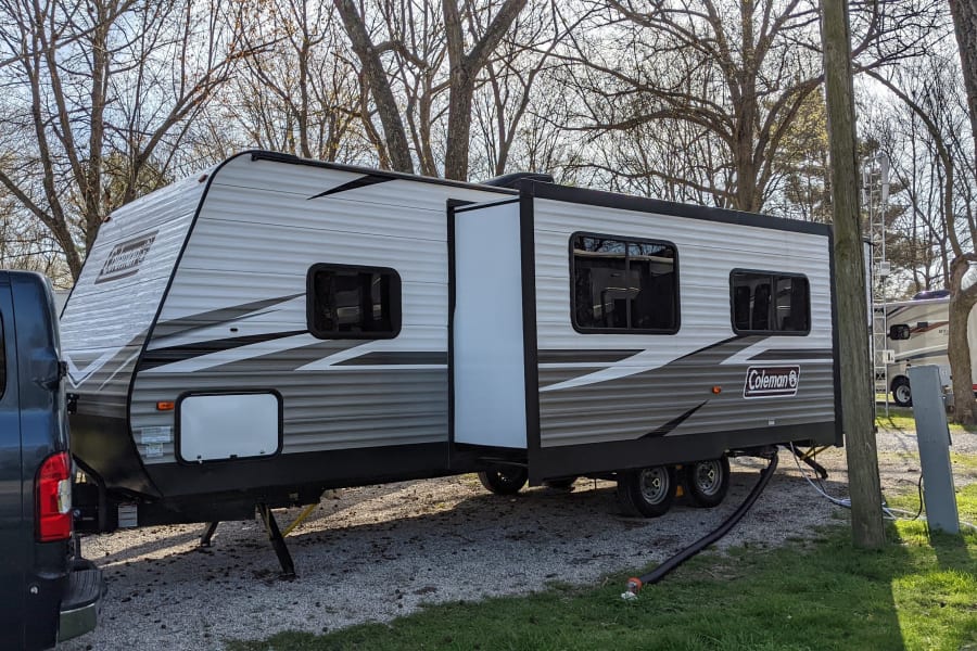 Experience an RV rental: a Coleman travel trailer parked with its slide-out extended at a sunny, gravel campsite.