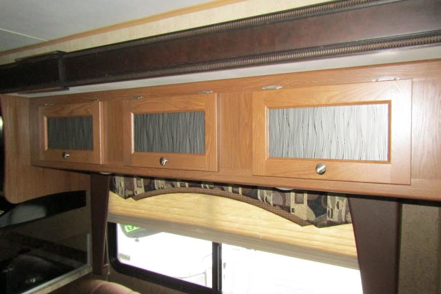 Inside an RV rental: wood overhead storage cabinets with glass inserts, window with shade, & seating.