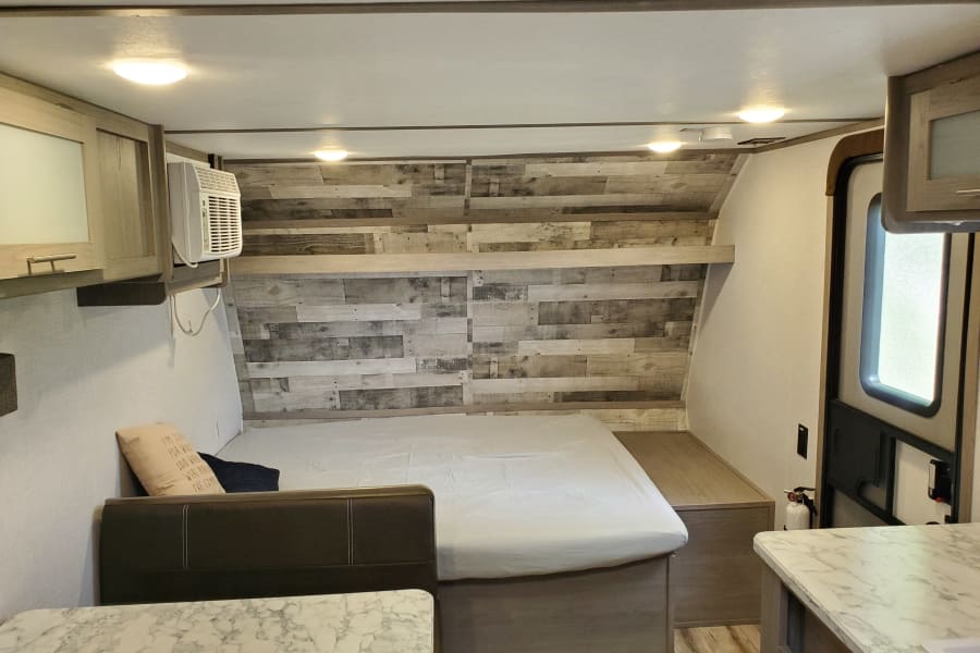 Inside a modern **rv rental**: a cozy bed with wood accent wall, overhead cabinets, AC, and marble countertop. Bright and functional interior.