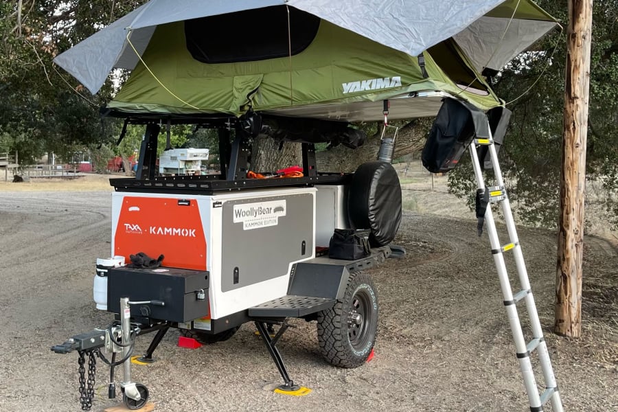 This compact RV rental camper trailer with a green Yakima rooftop tent and ladder is set up for outdoor camping adventures.