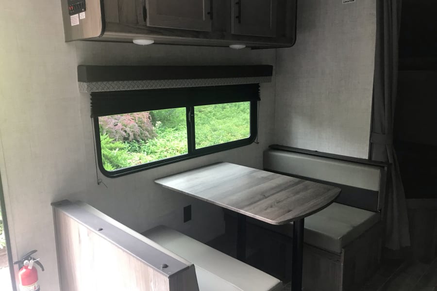 This RV rental features a cozy dinette with light benches, a wood-look table, and a large window showing lush greenery.