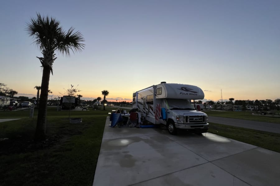 "Four Winds" RV rental at a palm-lined campground at sunset. People relax outside.