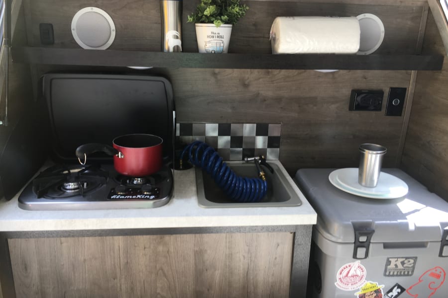 RV rental's compact kitchen setup featuring a gas stove, sink with pull-out hose, shelves, and a K2 cooler.