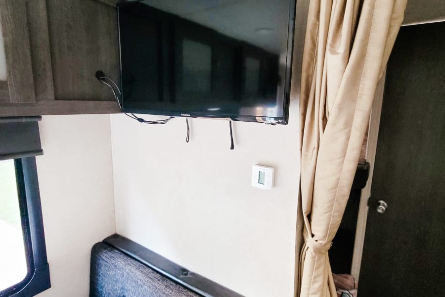 Compact RV rental interior, showcasing a mounted TV, dinette seating, overhead storage, and privacy curtain. Ideal for travel.