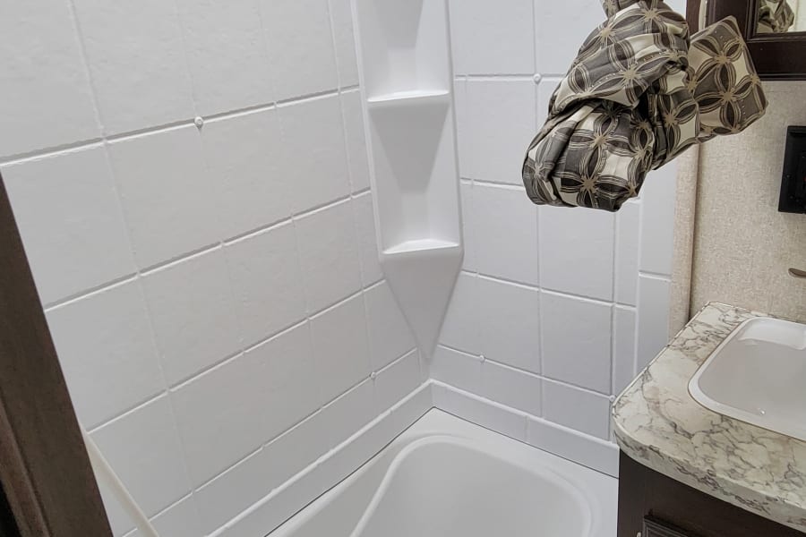 Clean white tiled shower and tub area with built-in shelves in an RV rental bathroom, featuring a decorative patterned curtain.