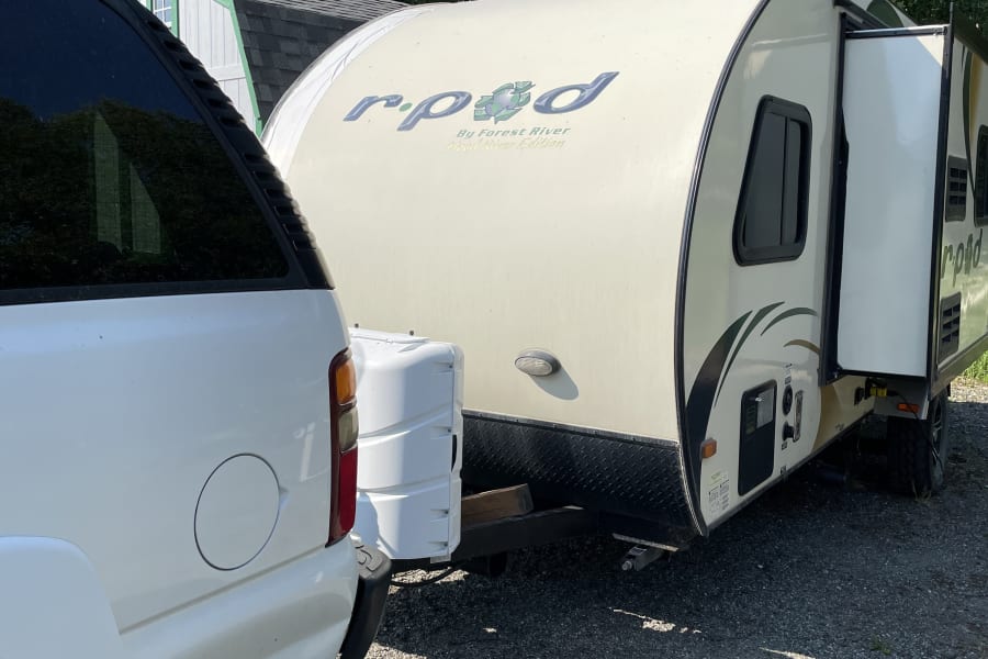 White SUV connected to a light-colored r-pod travel trailer, parked on gravel. Ideal for your next rv rental.