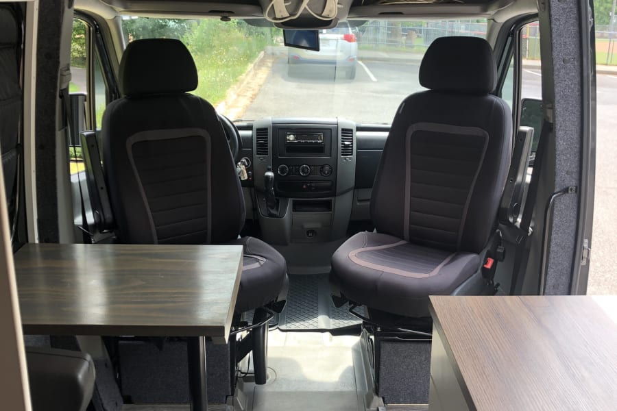 RV rental camper van interior showing front cabin seats, dashboard, a table, and overhead storage, with a wooden ceiling.