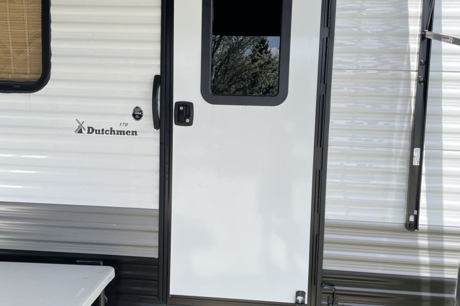 Experience an RV rental! This image shows the RV entrance with steps, a folding table, and outdoor mat, ready for camping.