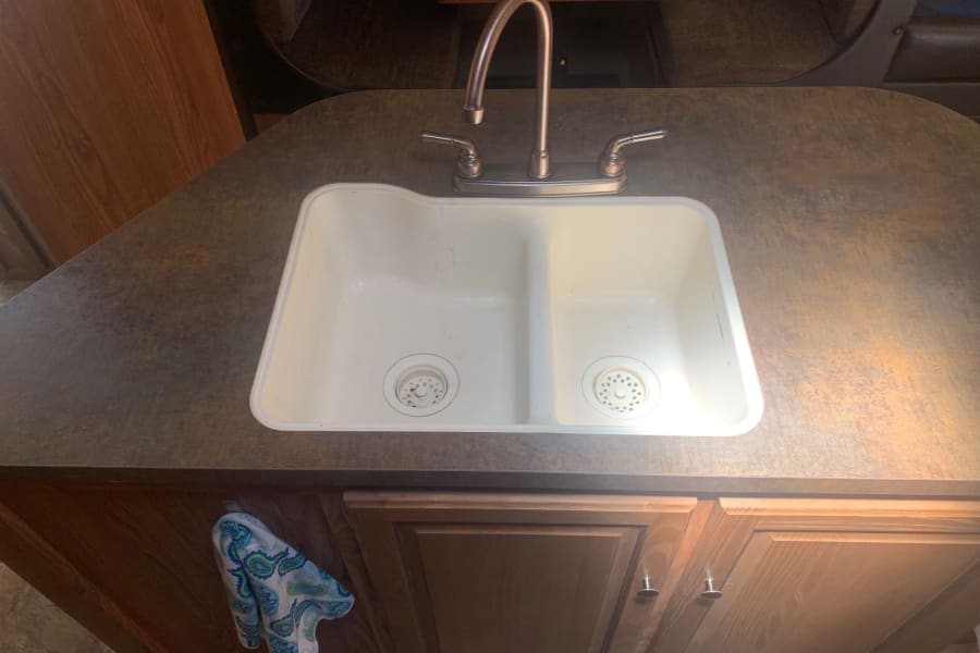 Clean double sink and bronze faucet in an RV rental kitchen, featuring a speckled countertop and wooden cabinets.