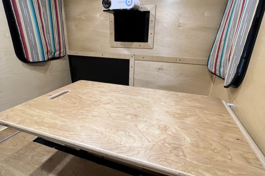 RV rental: Light wood interior with projector, striped window shades, and a large convertible table/bed platform.