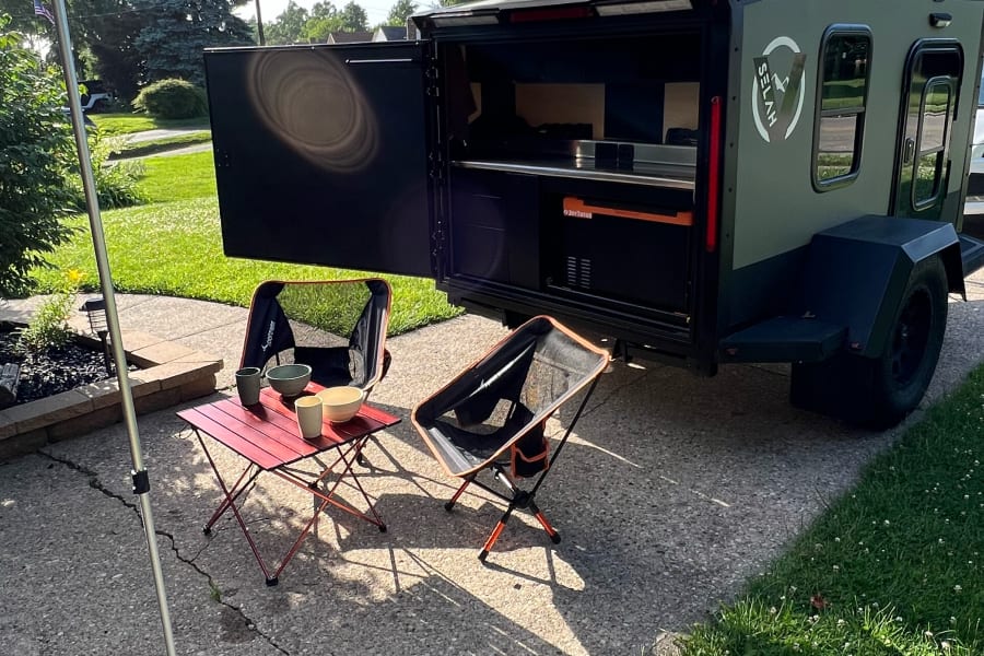 An RV rental: a dark green travel trailer with its back hatch open to reveal a kitchen. An awning extends, with two chairs & a table set up.