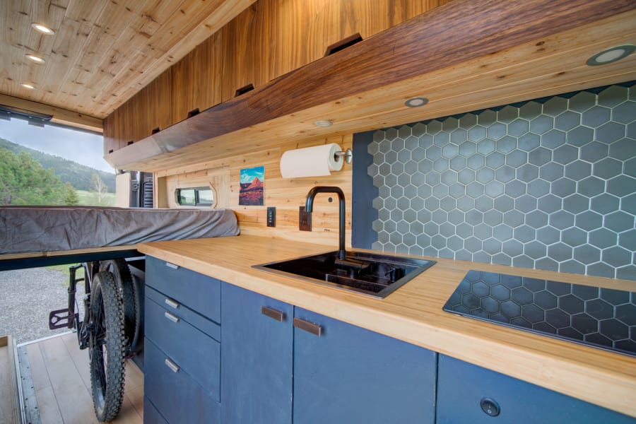 Modern RV rental interior: kitchen with hexagonal tiles, wood counters, bike storage, and mountain views.