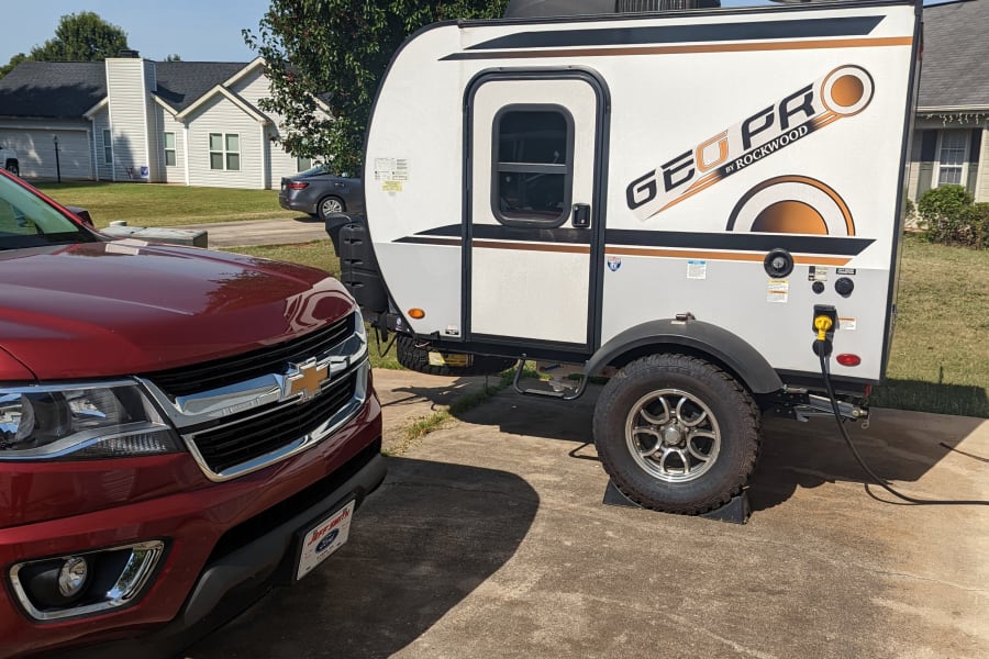 Red Chevy truck hitched to a white Geo Pro travel trailer, likely an RV rental, parked on a driveway on a sunny day.