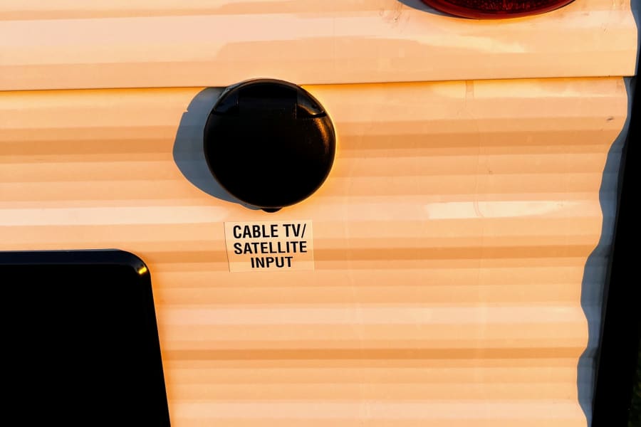 Side of an RV rental: a red marker light and a black cap labeled 'CABLE TV/SATELLITE INPUT' for essential camper hookups.