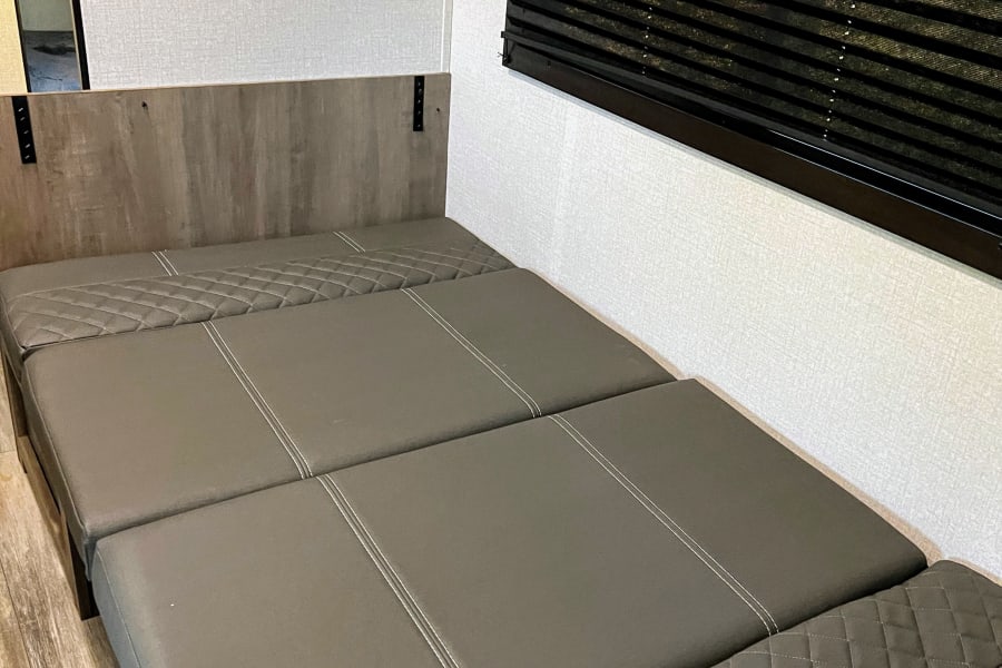 Modern rv rental interior showing a comfortable, convertible gray sofa bed with quilted sections, textured walls, and blinds.