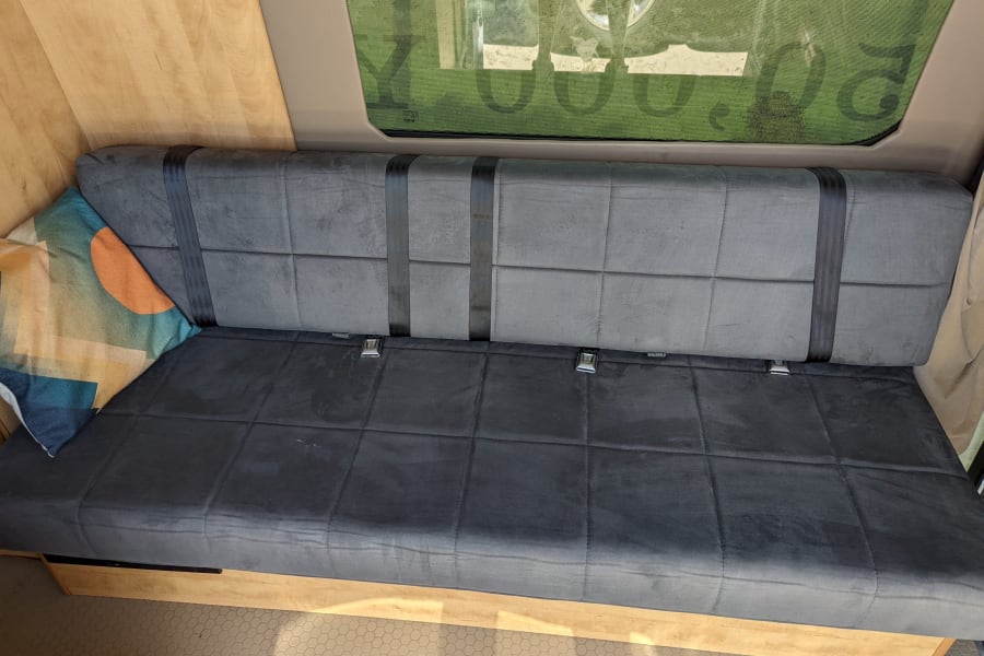 RV rental interior shows a gray bench seat with black seatbelts, a patterned pillow, and wooden walls next to a window.