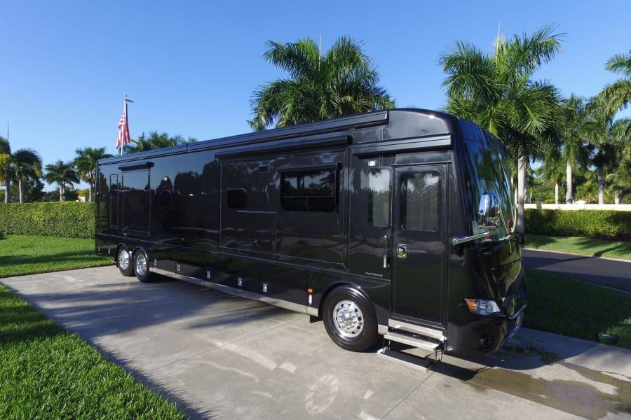 Sleek black luxury motorcoach RV rental parked on a sunny driveway, surrounded by lush palm trees & an American flag.